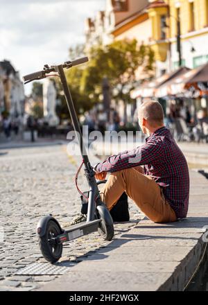 Jeune homme dans un pantalon décontracté assis sur le trottoir à côté de son scooter électrique garée, le fond flou de la ville Banque D'Images