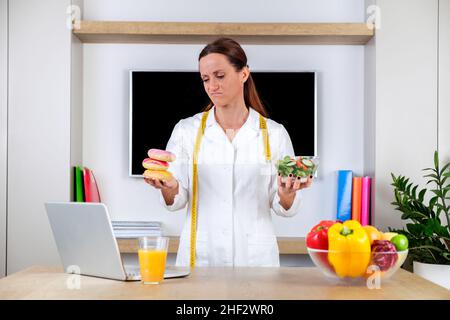 Jeune femme portant un uniforme nutritionniste tenant une salade et des beignets avec des émotions tristes dans une salle de bureau moderne et lumineuse.Choisir entre des aliments sains Banque D'Images