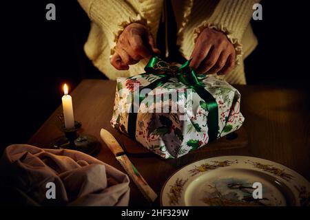 Une femme délie le ruban sur la boîte cadeau d'un gâteau de Noël allongé sur une table en bois avec une bougie en feu. Banque D'Images
