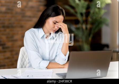 Jeune adulte fatigué triste surtravaillé femme d'affaires caucasienne, gestionnaire, secrétaire, épuisé en travaillant avec un ordinateur portable, fatigué de s'asseoir dans le bureau, frotte le pont de son nez, ferme les yeux Banque D'Images