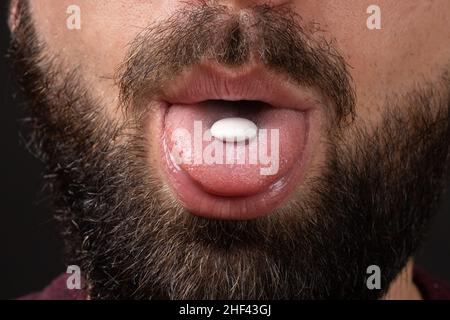 comprimé rond blanc sur la langue de la bouche ouverte de l'homme barbu avec la moustache foncée sur fond noir, prendre des vitamines, analgésique Banque D'Images