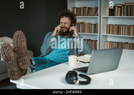 Le modèle masculin pose avec les pieds sur la table et le smartphone dans sa main.il y a ordinateur portable, Mug of Tea, Journal et écouteurs sur le bureau blanc .Arrière-plan des étagères.Gros plan .Photo de haute qualité Banque D'Images