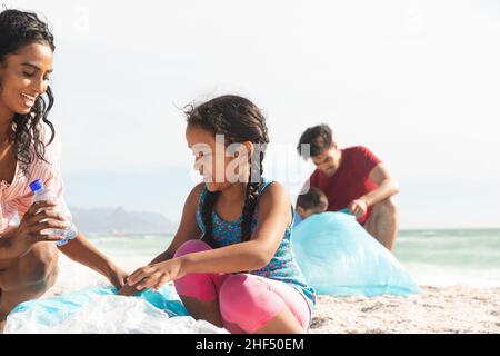 Bonne mère et fille biraciale collectant les déchets de bouteille en plastique dans le sac à la plage le jour ensoleillé Banque D'Images