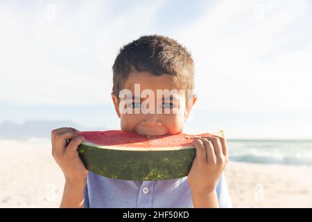 Portrait d'un adorable garçon biracial mangeant une grande tranche de pastèque fraîche à la plage le jour ensoleillé Banque D'Images