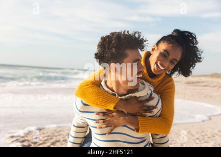Joyeux biracial homme donnant le pigeyback à la petite amie à la plage contre le ciel le jour ensoleillé Banque D'Images
