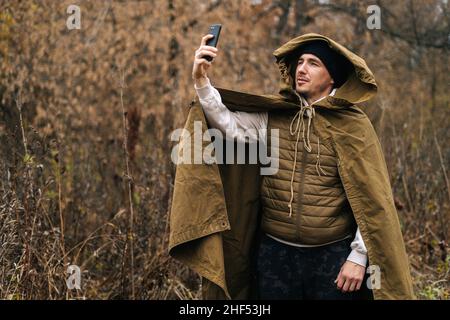 Portrait d'un homme survivaliste portant un imperméable à la tente à la recherche d'un signal de réception, à l'émission d'un appel vidéo ou à l'enregistrement d'un message vidéo sur un smartphone. Banque D'Images