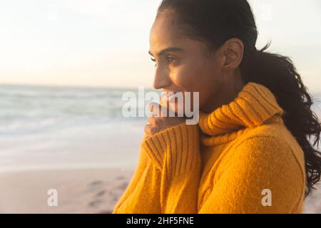 Jeune femme biraciale réfléchie portant un chandail jaune et regardant la plage pendant le coucher du soleil Banque D'Images