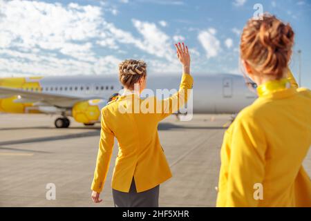 Vue arrière de deux hôtesses regardant l'avion et faisant bonjour geste tout en se tenant à l'extérieur de l'aéroport Banque D'Images