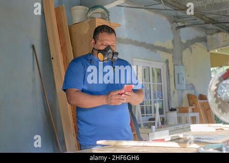 homme latin avec masque noir vérifiant le téléphone cellulaire, dans l'atelier de menuiserie Banque D'Images