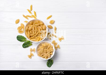 Vue de dessus sur une variété de pâtes italiennes crues dans des bols blancs avec des feuilles d'épinards verts frais sur fond de bois blanc Banque D'Images