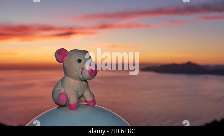 Grèce, Iles grecques, Iles Ioniennes, Corfou, côte ouest,Pentati, coucher de soleil, cochon farci assis sur une lampe sphérique blanche, en arrière-plan coucher de soleil sur la mer, ciel bleu avec des nuages rouges Banque D'Images