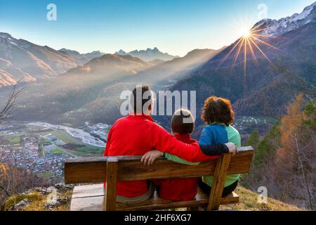 Famille de trois personnes (mère, père et fille) assise sur un banc en automne, en regardant le coucher du soleil, Agordo, province de Belluno, Vénétie, Italie Banque D'Images