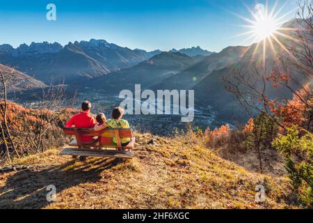 Famille de trois personnes (mère, père et fille) assise sur un banc en automne, en regardant le coucher du soleil, Agordo, province de Belluno, Vénétie, Italie Banque D'Images