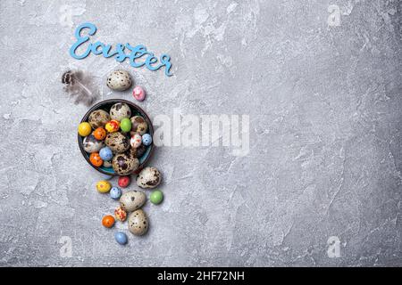 Vue de dessus de petits œufs de caille et de bonbons colorés dans un bol pour Pâques sur fond de béton gris avec espace de copie Banque D'Images