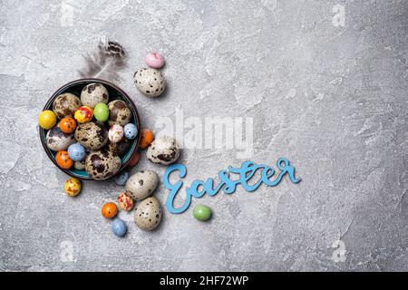 Vue de dessus de petits œufs de caille et de bonbons colorés dans un bol pour Pâques sur fond de béton gris Banque D'Images