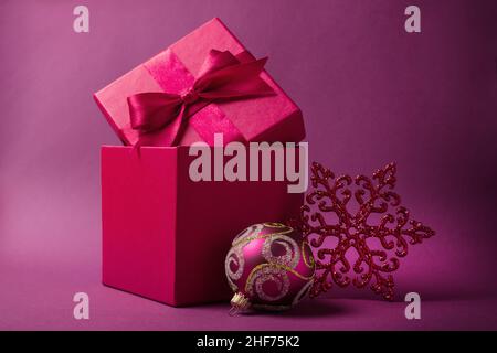 Magnifique boîte cadeau carrée rouge avec noeud satiné brillant pour le présent et boule pourpre, flocon de neige pour arbre de noël sur fond rose Banque D'Images