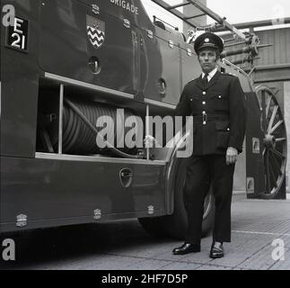 1960s, historique, un officier de station de la brigade des pompiers de Londres debout dans son uniforme par un moteur d'incendie Dennis à la caserne des pompiers de Lewisham, sud-est de Londres, Angleterre, Royaume-Uni.Les frères Dennis, John et Raymond, ont commencé à fabriquer et à vendre des bicyclettes en 1895, avant de progresser vers les voitures et les véhicules commerciaux et le légendaire moteur d'incendie. Banque D'Images