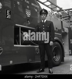 1960s, historique, un officier de station de la brigade des pompiers de Londres debout dans son uniforme par un moteur d'incendie Dennis à la caserne des pompiers de Lewisham, sud-est de Londres, Angleterre, Royaume-Uni.Les frères Dennis, John et Raymond, ont commencé à fabriquer et à vendre des bicyclettes en 1895, avant de progresser vers les voitures et les véhicules commerciaux et le légendaire moteur d'incendie. Banque D'Images