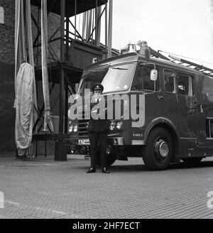 1960s, historique, un officier de station de la brigade des pompiers de Londres debout dans son uniforme par un moteur d'incendie Dennis à la caserne des pompiers de Lewisham, sud-est de Londres, Angleterre, Royaume-Uni.Les frères Dennis, John et Raymond, ont commencé à fabriquer et à vendre des bicyclettes en 1895, avant de progresser vers les voitures et les véhicules commerciaux et le légendaire moteur d'incendie. Banque D'Images