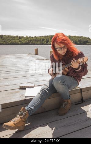 Adolescente en lunettes avec cheveux rouges joue ukulele assis sur une jetée en bois, vertical photo extérieure Banque D'Images