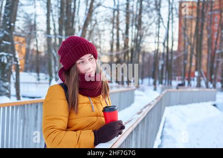 une jeune femme marche un hiver glacial.Une veste jaune vif, un chapeau bordeaux et une tasse en papier avec une boisson dans les mains.Copier l'espace Banque D'Images