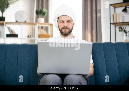 Vue rapprochée de l'avant d'un beau jeune homme caucasien barbu, assis sur un canapé bleu à la maison avec une serviette sur sa tête et des taches d'hydrogel rose sous les yeux, travaillant sur un ordinateur portable.Concentrez-vous sur l'ordinateur portable Banque D'Images