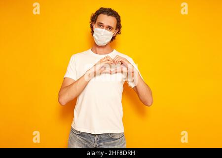 jeune homme en masque médical protecteur, vêtements décontractés, isolé sur fond jaune, se replie les mains en forme de coeur sur sa poitrine comme signe de gratitude à l'amour. concept de personnes, sécurité, santé.Photo de haute qualité Banque D'Images