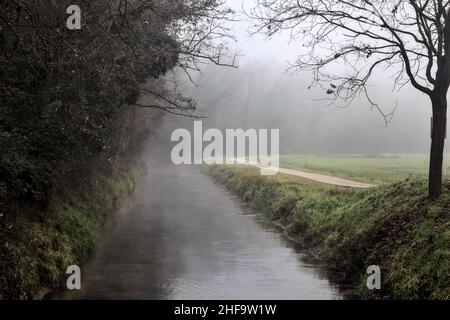 Ruisseau d'eau qui borde une forêt avec un chemin de terre par son bord dans la campagne italienne lors d'une journée brumeuse en hiver Banque D'Images