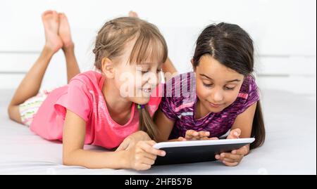 Deux petites filles mignonnes utilisent une tablette numérique et sourient lorsqu'elles sont allongées sur le lit dans la chambre des enfants Banque D'Images