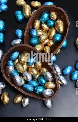 Pile de différentes tailles d'oeufs de pâques au chocolat enveloppés d'une feuille colorée en bleu, argent et or avec deux moitiés d'un gros oeuf de chocolat. Banque D'Images