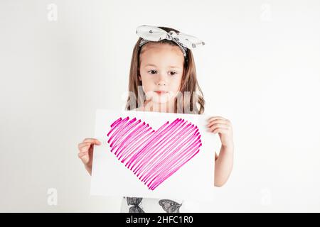 Portrait de petite fille réfléchie avec de longs cheveux tenant dans les mains feuille de papier avec coeur rose sur fond blanc.Félicitations pour votre famille Banque D'Images