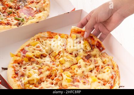 main d'une personne prenant un morceau de pizza avec du fromage, du jambon et de l'ananas, ingrédients tropicaux dans la restauration rapide, glucides dans emballage carton Banque D'Images