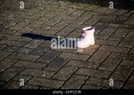 Une chaussure wellington rose pour bébé/petit enfant est jetée sur une passerelle pavée.La chaussure projette de longues ombres sous le soleil de la fin de l'après-midi. Banque D'Images