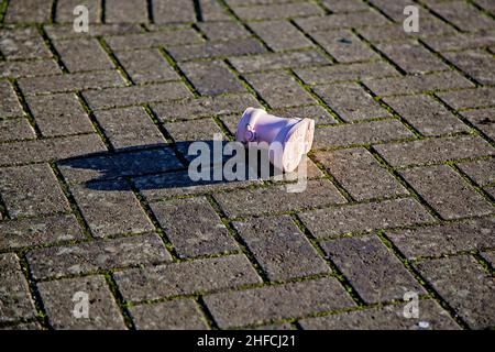 Une chaussure wellington rose pour bébé/petit enfant est jetée sur une passerelle pavée.La chaussure projette de longues ombres sous le soleil de la fin de l'après-midi. Banque D'Images