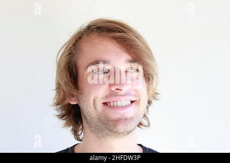 Jeune homme aux yeux verts souriant en franchise.Jeune homme aux longs cheveux blonds souriant isolé candidement sur fond blanc.Expressions du visage. Banque D'Images