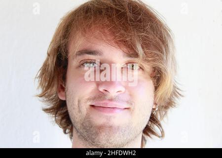 Jeune homme aux yeux verts souriant en franchise.Jeune homme aux longs cheveux blonds souriant isolé candidement sur fond blanc.Expressions du visage. Banque D'Images