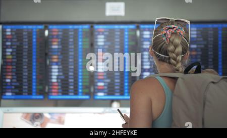 Femme caucasienne à l'aéroport de Suvarnabhumi portant un masque médical de protection. Tableau d'information de vol. Utilisez un smartphone mobile. Virus de la santé protéger l'épidémie du coronavirus sras-cov-2 covid-19 2019-ncov. Banque D'Images