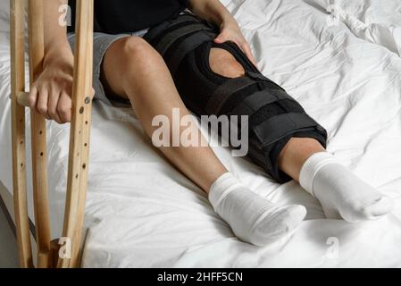 Une patiente portant une orthèse assise sur un lit tenant des béquilles après une intervention chirurgicale sur une jambe cassée.Réadaptation de la femme blessée à la maison.Mise au point sélective Banque D'Images