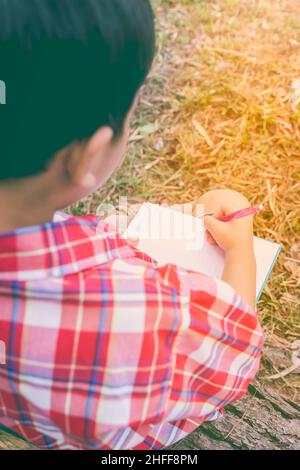 Vue arrière.Enfant utilisez le stylo pour écrire sur le livre dans le parc.À l'extérieur, en journée, en plein soleil.Les enfants lisent et étudient, concept d'éducation.Chaud t Banque D'Images