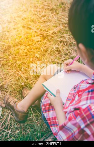 Vue arrière.Enfant utilisez le stylo pour écrire sur le livre dans le parc.À l'extérieur, en journée, en plein soleil.Les enfants lisent et étudient, concept d'éducation.Chaud t Banque D'Images