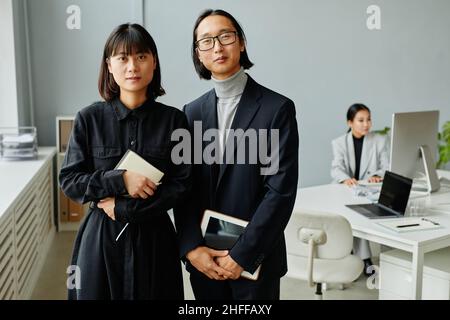 Portrait à la taille de deux entrepreneurs asiatiques regardant l'appareil photo tout en étant debout au bureau Banque D'Images