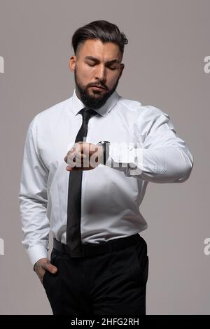 Homme hispanique barbu dans un gilet sombre et une chemise blanche avec cravate regardant l'appareil photo avec les mains dans les poches en studio Banque D'Images