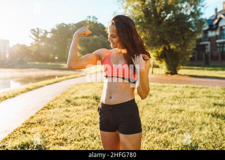 jeune femme athlétique aux cheveux foncés dans les vêtements de sport en flexion de son biceps dans le parc dans la journée ensoleillée d'été. Banque D'Images