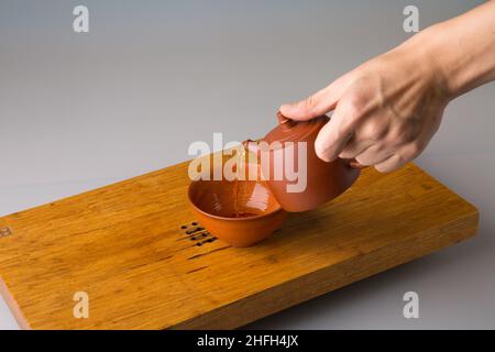 La main d'un homme verse du thé pu erh fraîchement préparé d'une théière en argile dans une tasse traditionnelle.Une tasse sur une table pour les cérémonies du thé. Banque D'Images