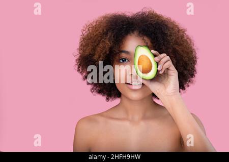 Portrait d'une belle jeune femme à la peau sombre avec des cheveux bouclés couvre un oeil avec la moitié d'un avocat regarde l'appareil photo et sourit sur un fond rose. Banque D'Images