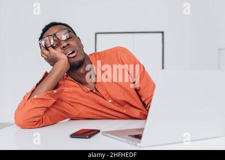 Un jeune homme afro-américain fatigué avec des lunettes dort à portée de main près d'un ordinateur portable à une table blanche dans une salle lumineuse vue rapprochée Banque D'Images