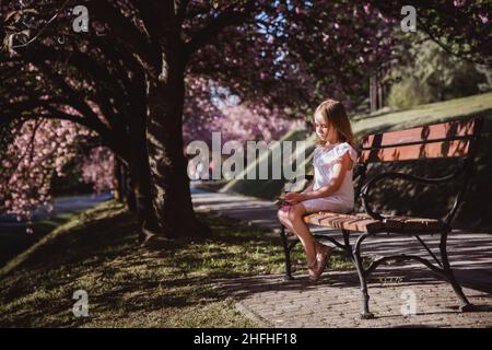 Adorable petite fille en robe blanche est assise sur un banc dans un jardin rose fleuri le beau jour de printemps Banque D'Images