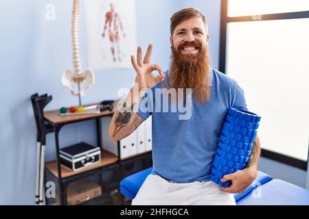 REDHEAD homme avec la longue barbe tenant le rouleau en mousse à la clinique médicale faisant signe ok avec les doigts, souriant sympathique gestant excellent symbole Banque D'Images