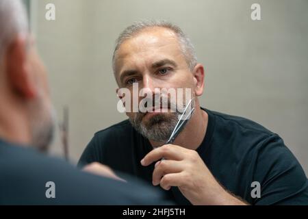 Portrait d'un beau homme d'âge moyen coupant sa barbe avec des ciseaux.Autosoins pendant l'isolement en quarantaine Banque D'Images