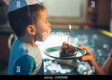 Gros plan d'un petit garçon soufflant les bougies sur le morceau de gâteau donné par maman.Mets sucrés.Fête des fêtes.Fête d'anniversaire. Banque D'Images